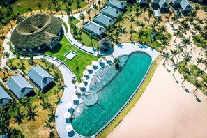 Vista aérea de un complejo turístico con una piscina central, varias cabañas y un edificio circular de techo de paja.