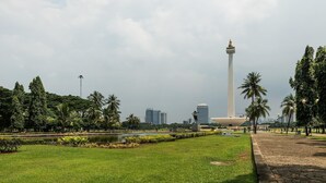 A park with a tall monument, a pond, and a cityscape in the background. - H! Hotel Senen (Jakarta)