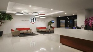 A modern reception area with a clean, minimalist design, featuring a reception desk, two sofas with red cushions, and a large potted plant. - H! Hotel Senen (Jakarta)