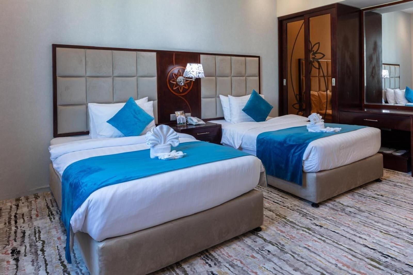 A hotel room with two beds, each with a blue blanket and white linens. There is a wooden headboard with a decorative panel and a wall-mounted lamp.