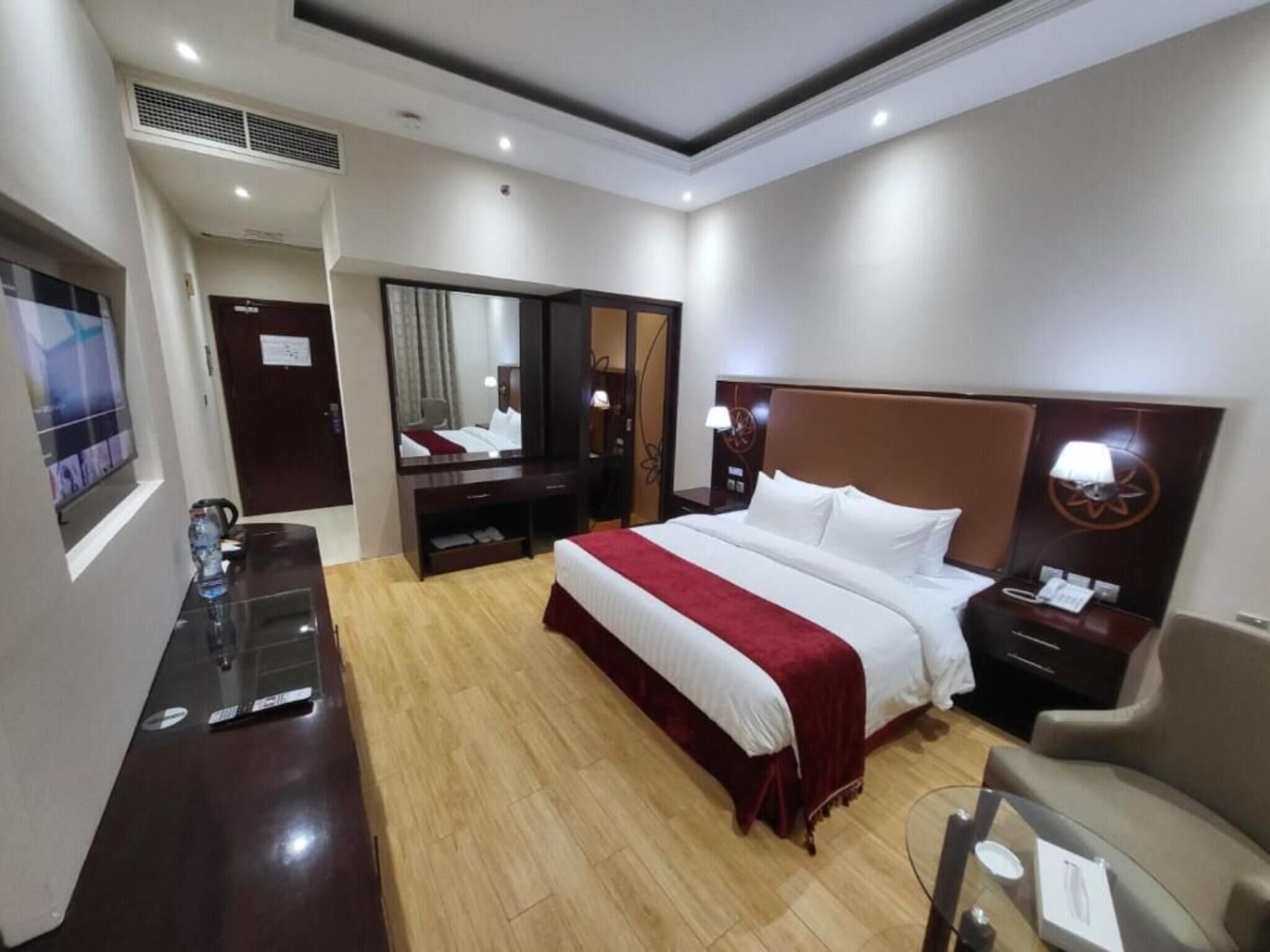 A hotel room with a large bed, a desk, a chair, a mirror, and a nightstand with a lamp.
