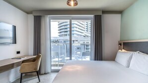 A hotel room with a large bed, a desk with a chair, a flat-screen TV, and a view of a modern building through the window. - Hotel Mistral St. Julian's (St. Julian's)