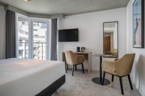 Quarto de hotel com cama, escrivaninha, cadeira, televisão e uma janela grande.