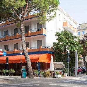A multi-story hotel with outdoor seating, a tree, and a clear sky. - Hotel Taormina (Jesolo)