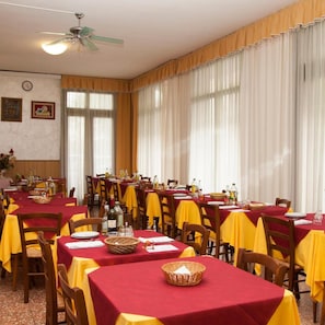 Zona pranzo con tavoli apparecchiati, sedie, ventilatore a soffitto e ampie finestre con tende.
