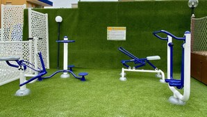 Une salle de sport en plein air, dotée d’appareils d’exercice installés sur une pelouse artificielle, entourée d’une clôture blanche et d’une haie verte.