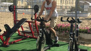 Une personne fait de l’exercice sur un vélo d’appartement, à l’abri, avec un banc et du matériel de musculation en arrière-plan.