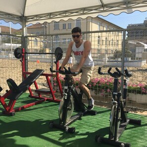 A person exercising on a stationary bike under a covered area with a bench and weightlifting equipment in the background. - Hotel Jumbo (Rimini)