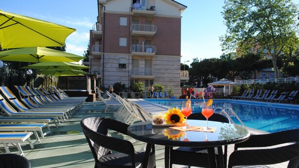 Un espace aménagé au bord de la piscine, avec des chaises longues, une table avec des boissons et un bâtiment en arrière-plan.