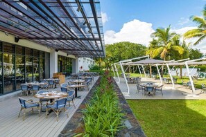 An outdoor dining area with tables and chairs, a covered walkway, and a view of palm trees. - Ocean's Creek (Balaclava)