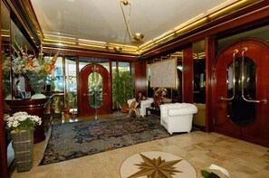 A lobby with a reception desk, a large mirror, a white sofa, and a patterned rug. - Hotel Beny (Jesolo)