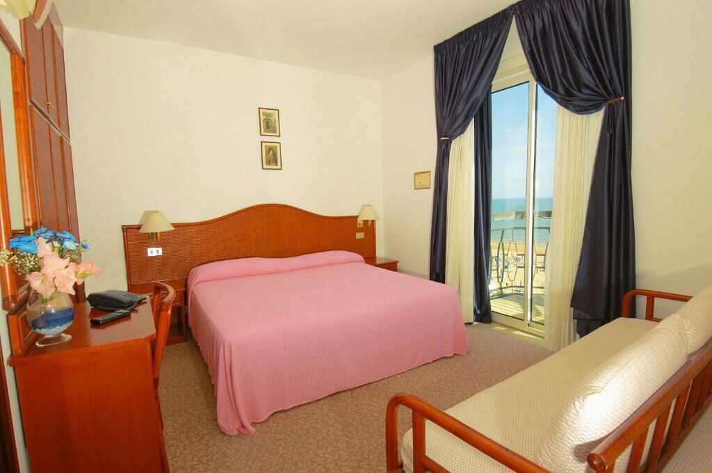 Een hotelkamer met een bed, een nachtkastje met een lamp, een stoel en uitzicht op zee door een groot raam.