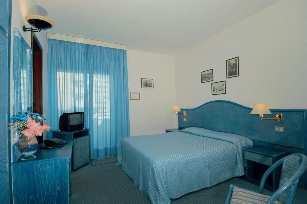 A hotel room with a bed, a desk with a television, a chair, and a lamp.