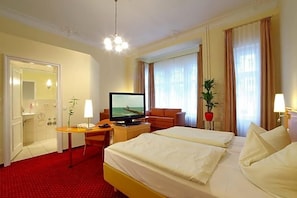 A hotel room with a bed, a desk with a television, a chair, a plant, and a bathroom with a mirror.