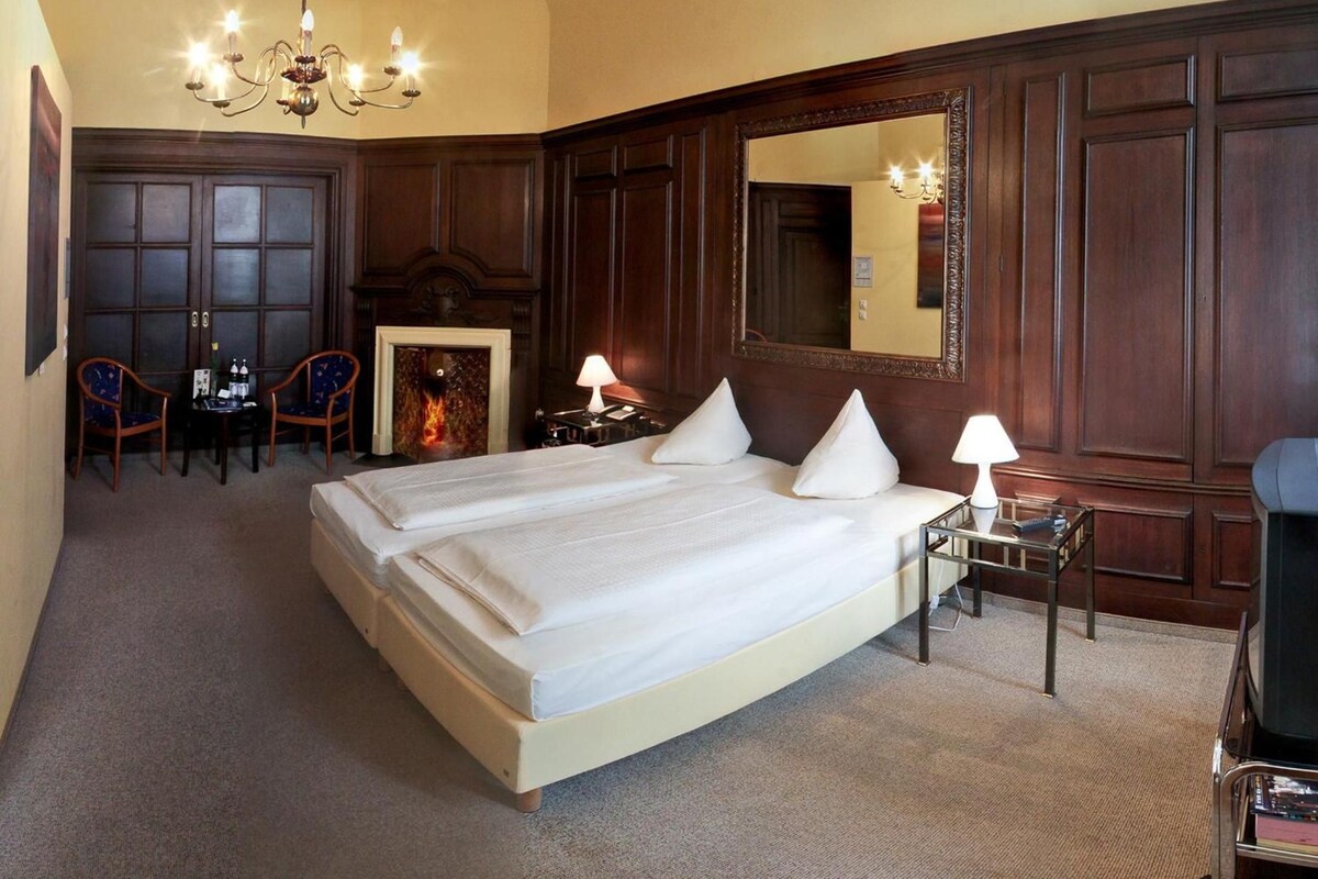 Une chambre d’hôtel avec un grand lit, une cheminée, un miroir, un bureau avec une lampe et une chaise.