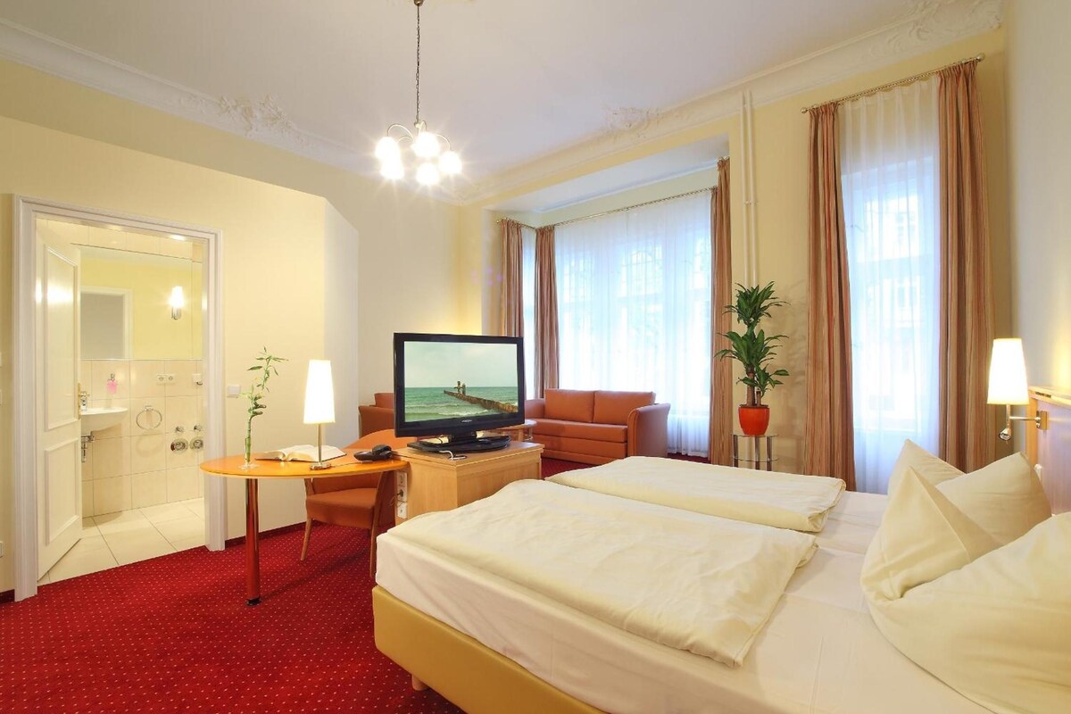 Une chambre d’hôtel comprenant un lit, un bureau avec une télévision, une chaise, une plante en pot et une salle de bain avec un miroir et un lavabo.