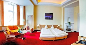 A hotel room with a bed, a sofa, a chair, a small table, a fireplace, and a painting on the wall.