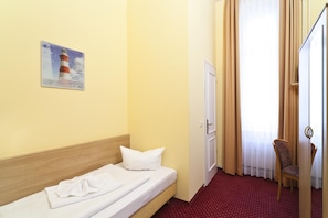 Une chambre d’hôtel avec un lit simple, une tête de lit en bois, une moquette rouge, une chaise et une porte avec des rideaux.