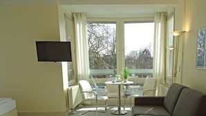 Un salon moderne avec un canapé, un téléviseur fixé au mur, une petite table ronde avec des chaises et de grandes fenêtres avec des rideaux.