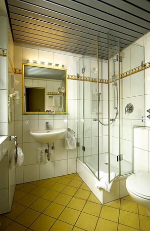 A bathroom with a glass-enclosed shower, a sink, a mirror with lights, and a toilet. - Hotel Ludwig Superior (Cologne)