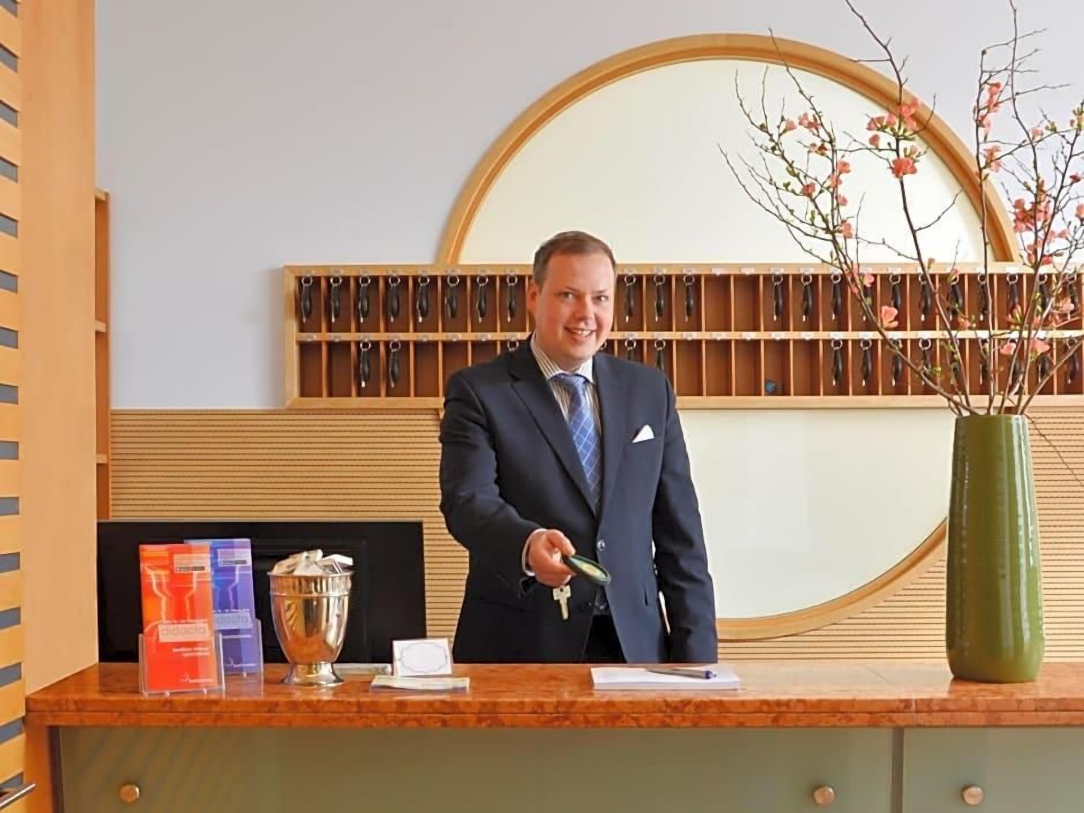 Un homme en costume, debout derrière un comptoir d’accueil, avec un miroir, des clés et un vase de fleurs.