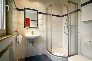 A bathroom with a glass-enclosed shower, a sink, and a mirror with a blue-tiled border. - Hotel Ludwig Superior (Cologne)