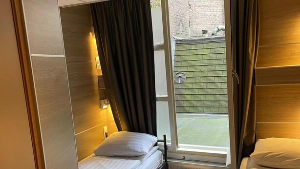 A compact hotel room with a single bed, a small table, and a window with a view of a brick wall and greenery. - Hotel Mevlana (Amsterdam)