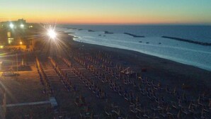 A beach with rows of umbrellas and chairs, a pier extending into the water, and buildings in the background. - Hotel Biagini (Rimini)