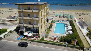 A multi-story hotel with a pool, beach view, and outdoor seating area. - Hotel Biagini (Rimini)