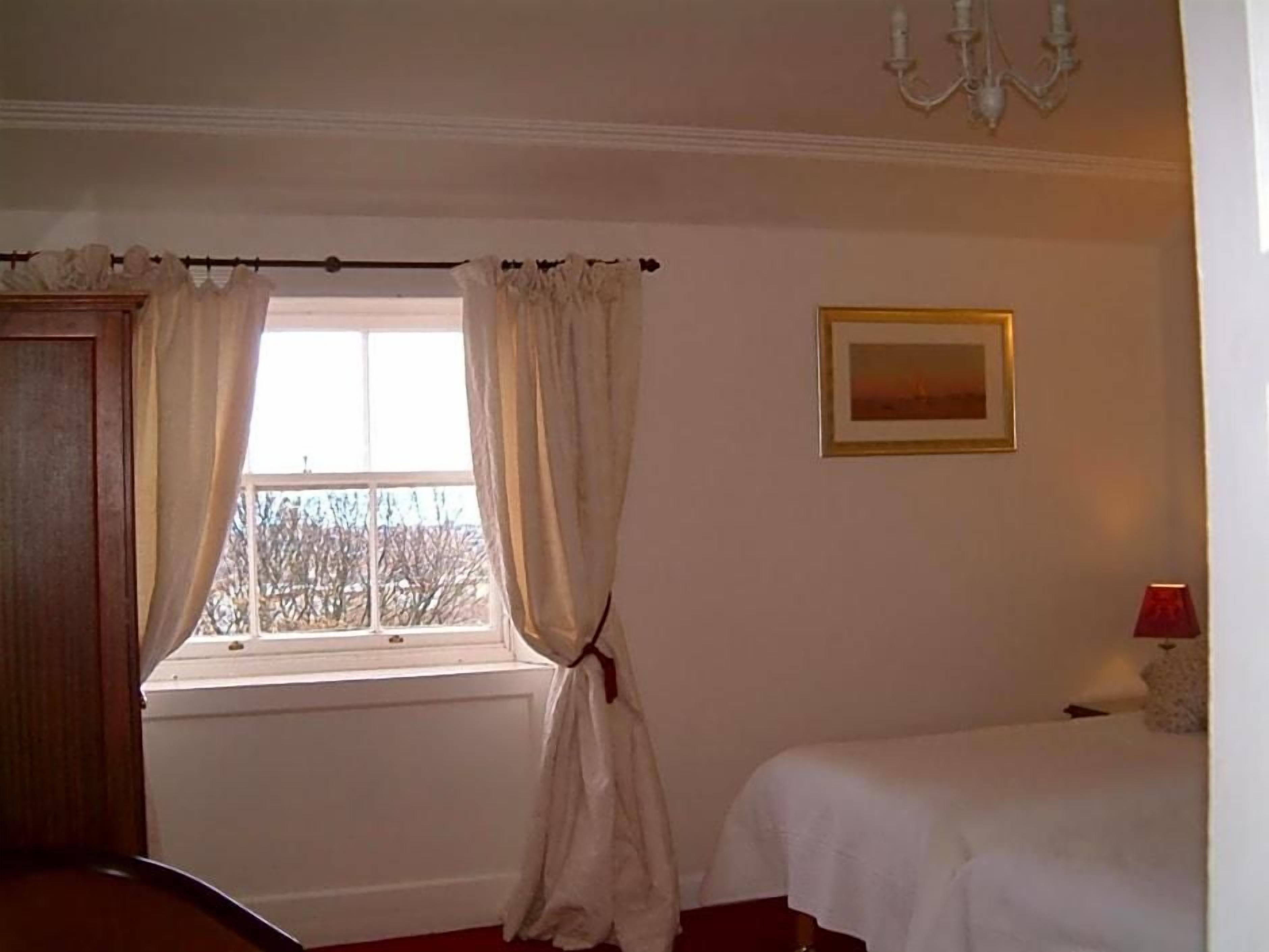 Una habitación con ventana, cama, mesita de noche, lámpara y un cuadro en la pared.