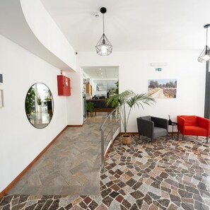Un hall d’entrée avec un sol carrelé en pierre, un comptoir d’accueil, une boîte rouge contenant du matériel de premiers secours, un miroir et un escalier menant à un autre espace.