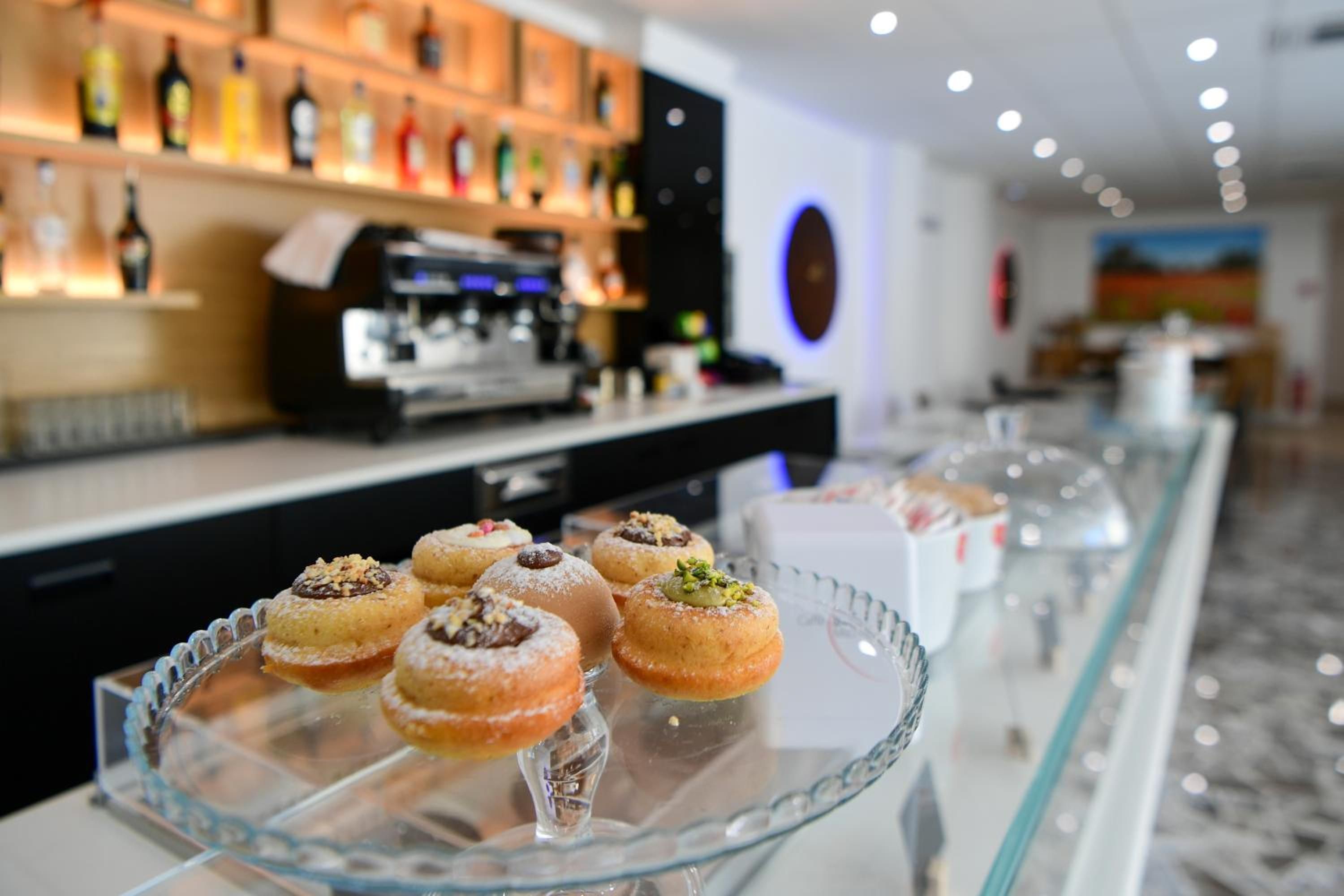 Esposizione di dolci con diversi pasticcini su un bancone in vetro, una macchina da caffè e un bar con bottiglie sullo sfondo.