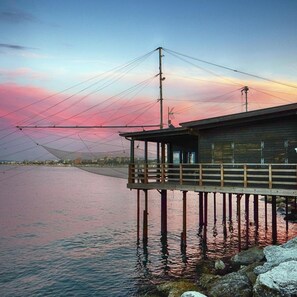 A pier extending into calm water with a wooden building at the end, supported by metal poles. - Hotel Ivano (Rimini)