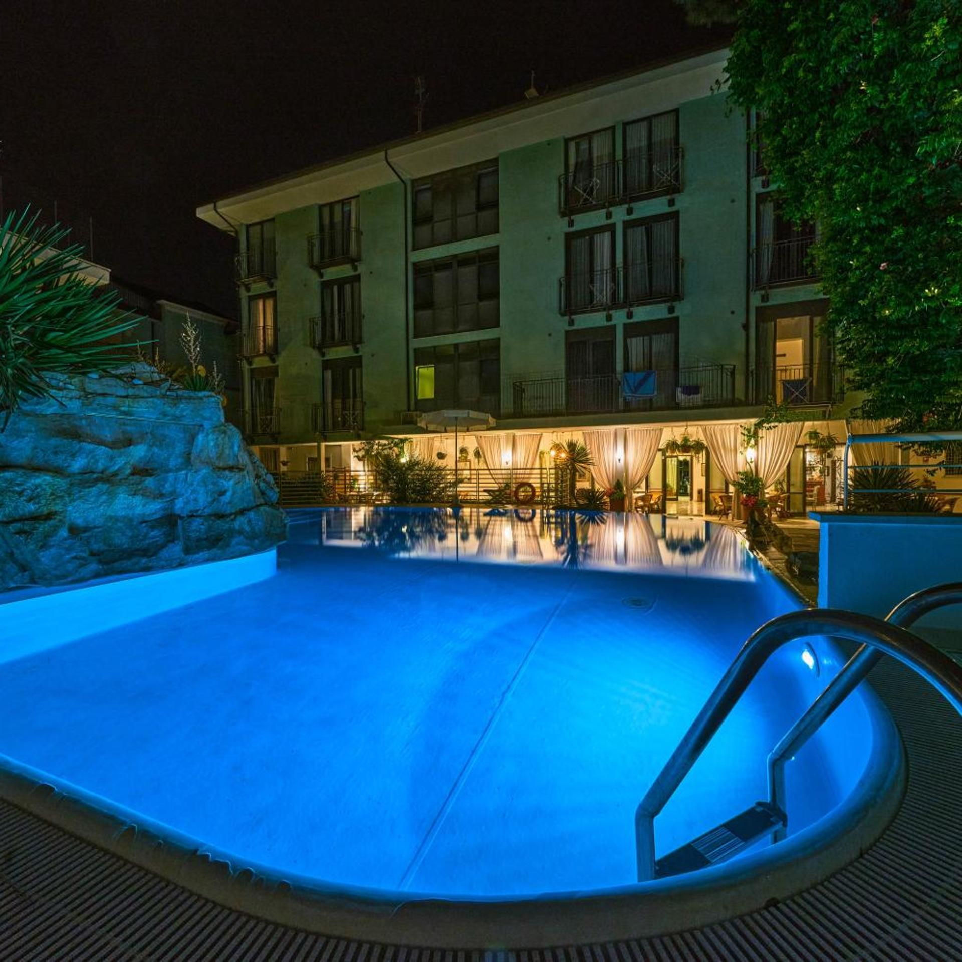 Vista nocturna d'un hotel amb piscina, mur de pedra i edifici amb diversos balcons.