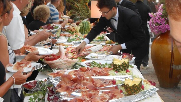 Een buffet met diverse gerechten, waaronder vlees en salades, buiten opgesteld met uitzicht op gebouwen op de achtergrond.