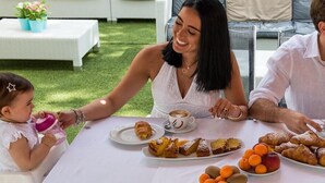 Une famille prend son repas à l’extérieur, autour d’une table où sont disposés des croissants, des pâtisseries et des fruits.