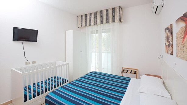 Une chambre à coucher avec un berceau, un lit, une télévision, un radiateur et une fenêtre avec des rideaux.