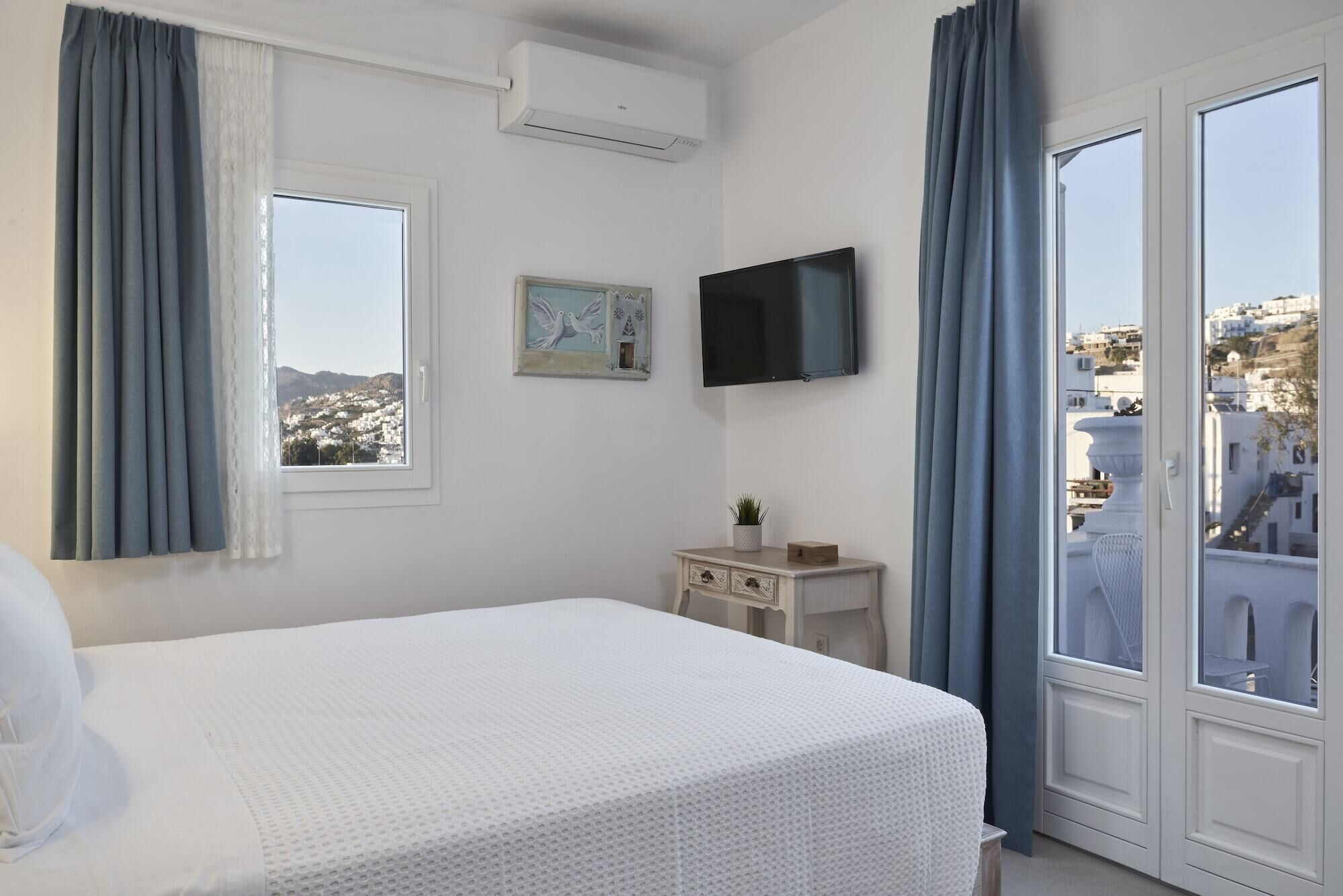 A hotel room with a bed, a TV, a small bedside table with a plant, and a view of the city.