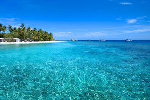 A clear turquoise sea with a sandy beach, palm trees, and a few boats in the distance. - Sandies Bathala Maldives (Bathala Island)