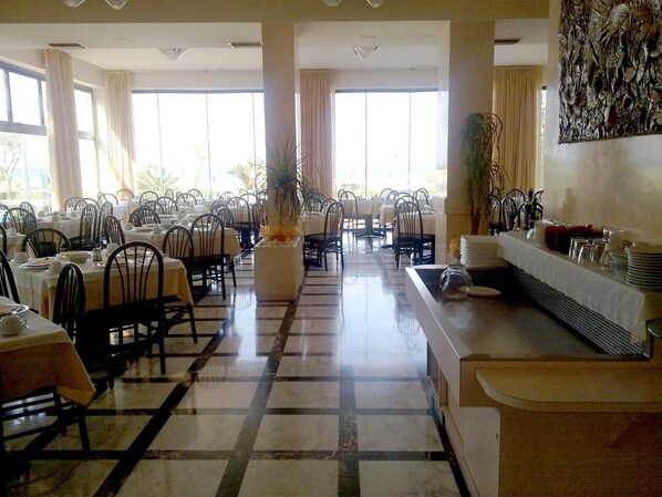 A spacious dining area with multiple tables and chairs, large windows, and a central decorative plant. - Cobalto (Rimini)