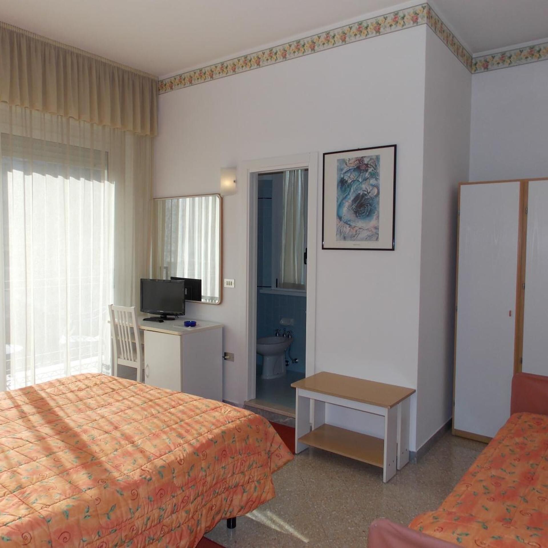 Habitación de hotel con dos camas, un escritorio con televisor, un baño y un cuadro en la pared.