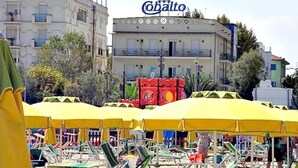 Ein Strand mit gelben Sonnenschirmen und Liegestühlen, ein Gebäude mit Balkonen und ein Schild mit der Aufschrift 'Cobalto'.