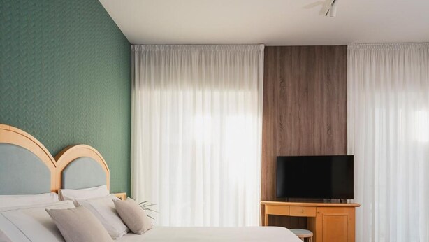 Hotellihuone, jossa on sänky, puinen TV-taso ja ikkuna, jossa on ohuet verhot.