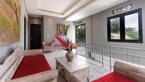 A spacious room with a large window, a wooden sofa set, a small table with a vase, and a painting on the wall. - Nawa Sakti Resort (Ubud)