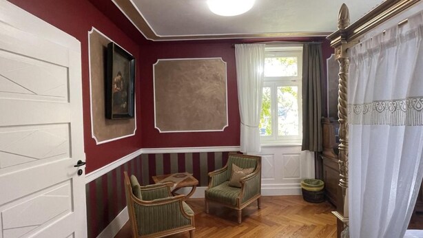 Une pièce avec un plancher en bois, deux fauteuils, une table, un grand tableau, une fenêtre avec des rideaux et une porte.