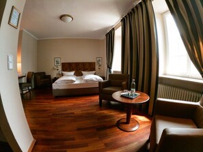 Hotelkamer met een bed, twee fauteuils, een klein rond tafeltje met een fles en glazen, houten vloeren en een raam met gordijnen.