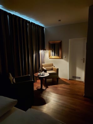 Quarto de hotel com sofá, poltrona, mesa pequena, abajur e espelho na parede.