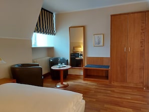 Quarto de hotel com cama, escrivaninha, cadeira, mesa pequena, guarda-roupa, espelho e janela com persiana riscada.