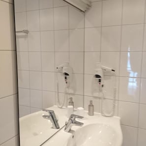 Een badkamer met witte betegelde muren, een wastafel met spiegel en twee haardroger bevestigd aan de muur.