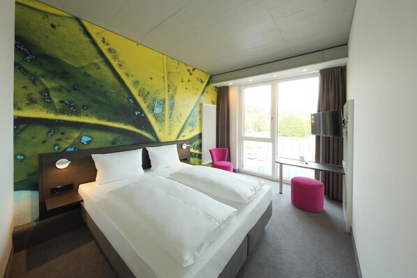 A hotel room with a large bed, a TV, a window with curtains, and a colorful wall mural. - Qubixx Stadtmittehotel (Schwäbisch Hall)